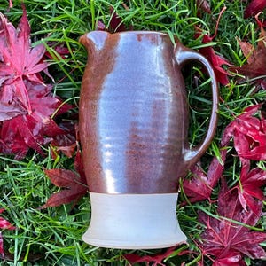 May include: A ceramic pitcher with a glossy, dark brown glazed upper body and a matte, light gray unglazed base. The vessel features a curved handle on the right side and a small pouring spout at the top rim, showcasing a two-tone finish.