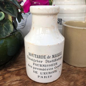 Puede incluir: Una botella de cerámica blanca vintage con texto negro "MOUTARDE de MAILLE". La botella tiene un cuello estrecho y una base más ancha. La botella está ligeramente envejecida con grietas y manchas visibles.