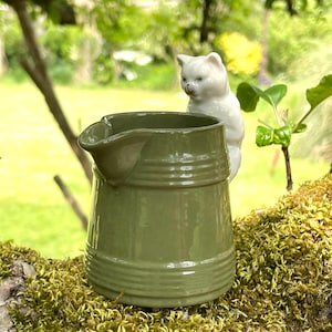 May include: A green ceramic pitcher with a spout and horizontal ridges, paired with a white cat figurine. The pitcher is a muted green color and sits on a mossy surface, with a blurred green background.