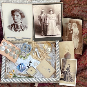 Puede incluir: Una caja abierta con fotografías antiguas y varios objetos antiguos. La caja contiene fotografías antiguas de personas, incluidos retratos de bodas. Otros artículos incluyen botones de nácar, encaje y un pequeño ancla plateada.