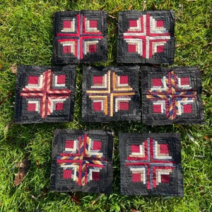 May include: Seven square fabric coasters with a log cabin design. Each coaster has a black background and a cross-shaped pattern in red, cream, and patterned fabrics. The coasters are arranged on green grass.