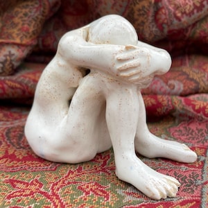 May include: A ceramic sculpture of a person in a seated, fetal position. The figure is white with brown speckles and has arms wrapped around the head. The sculpture is on a patterned red, green, and brown surface.