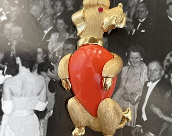 May include: Gold-coloured poodle brooch with a red centre. The poodle has a textured finish, a red rhinestone eye, and a gold collar. The brooch is set against a black and white background.
