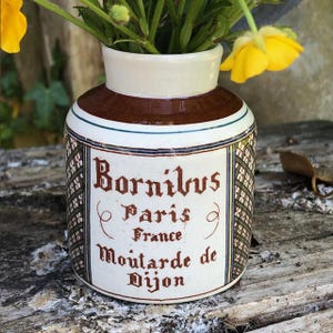 Puede incluir: Un tarro de cerámica vintage con el texto "Bornibus Paris France Moutarde de Dijon" en letras marrones. El tarro tiene una base blanca, una parte superior marrón y un patrón decorativo. Hay flores amarillas en el tarro.