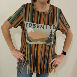 May include: A short-sleeved t-shirt with a vertical striped pattern in shades of brown, orange, and teal. The shirt features a graphic on the front with the text "YOSEMITE CALIFORNIA" and a mountain and tree design. The bottom hem has fringe.