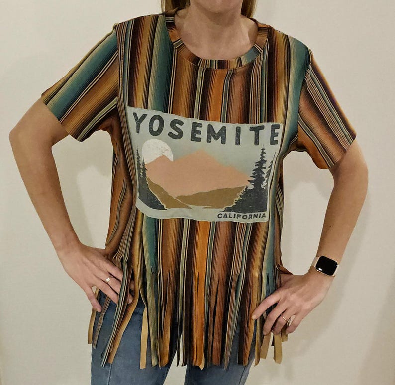 May include: A short-sleeved t-shirt with a vertical striped pattern in shades of brown, orange, and teal. The shirt features a graphic on the front with the text "YOSEMITE CALIFORNIA" and a mountain and tree design. The bottom hem has fringe.