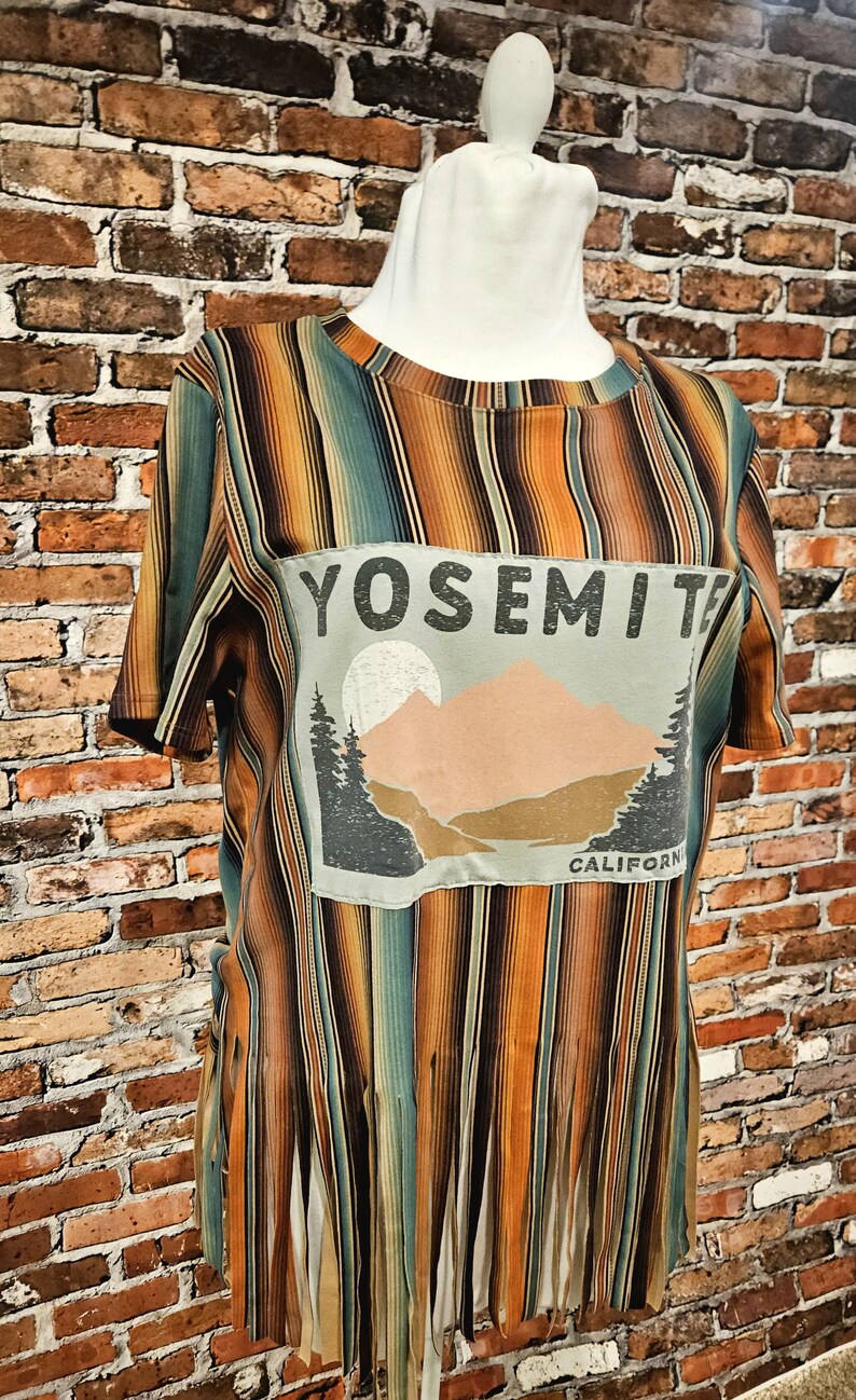 May include: A short-sleeved shirt with vertical stripes in shades of orange, brown, and teal. The shirt features a graphic of a mountain scene with the text "YOSEMITE CALIFORNIA". The bottom hem has a fringed design.