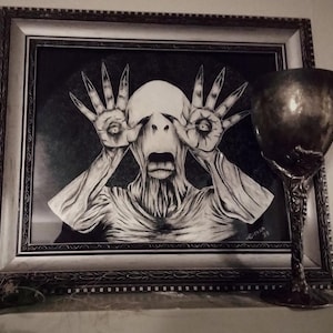 May include: A framed black and white artwork depicts a pale, humanoid figure with hands over its eyes and an open mouth. A decorative silver goblet stands to the right of the artwork, adding a gothic touch.