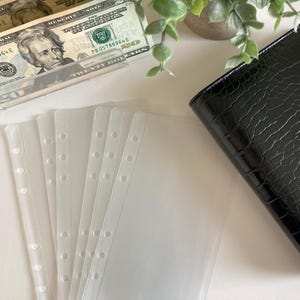 May include: Five clear plastic page protectors with six holes down the side for a binder. The protectors are lying on a white surface.
