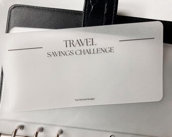 May include: A clear plastic card with the text "TRAVEL SAVINGS CHALLENGE" in a black font. The card is inside a black leather binder. The text "The Refined Budget" is at the bottom of the card.