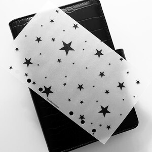 May include: A black, textured planner with a star-patterned overlay. The overlay is white with black stars of varying sizes. The text "The Refined Budget" is visible on the overlay. The planner is partially open.
