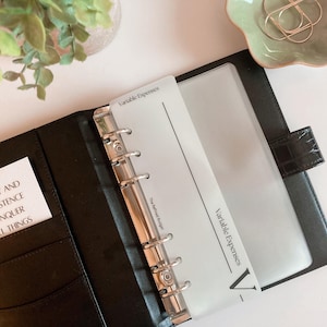 Könnte beinhalten: Schwarzer Lederordner mit silbernen Metallringen, geöffnet, um eine transparente Kunststoffhülle und einen weißen Trenner mit der Aufschrift "Variable Expenses" zu zeigen. Eine kleine Karte mit Text ist in einer Tasche sichtbar.