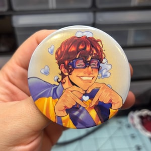 Puede incluir: Un botón redondo con una ilustración de una persona sonriente con cabello rojo, gafas azules y un atuendo azul y amarillo. La persona está haciendo una forma de corazón con sus manos, con pequeños corazones azules a su alrededor.