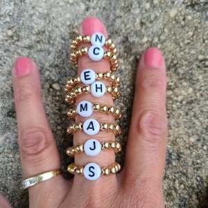 May include: A hand wearing several gold beaded rings with white lettered beads spelling out the names: N C, E H, M A, J, and S. The rings are on a finger with pink nail polish. A gold ring with diamonds is on the ring finger.