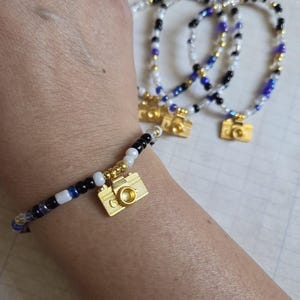 May include: Three beaded bracelets with gold camera charms. The bracelets are black, white, and blue.