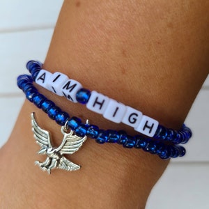 May include: Two blue beaded bracelets with a silver eagle charm. The bracelets spell out "AIM HIGH" in white letters.