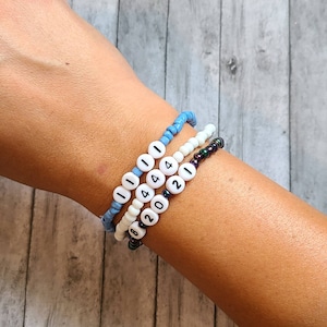 Puede incluir: Un conjunto de tres pulseras de cuentas con cuentas blancas y números negros. Las pulseras deletrean "1114442021".