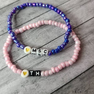 Puede incluir: Dos pulseras de cuentas. Una es rosa con cuentas de letras blancas que deletrean "OTH" y un corazón. La otra es azul con cuentas de letras blancas que deletrean "MSC" y un corazón. Las pulseras están sobre una superficie de madera gris.