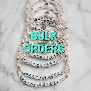 May include: A collection of white and pink beaded bracelets with gold accents. Each bracelet features a different word spelled out in black letters. The words include "Trayonna", "Pray For It", "Barbie Girl", "Self Reflect", "Goofy Goober", and "Main Squeeze". The bracelets are stacked on top of each other and the text "Bulk Orders" is written in large, turquoise letters in the center of the image.