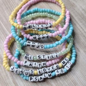 Könnte beinhalten: Ein Set aus bunten Perlenarmbändern mit Wörtern wie "Believe", "Hope", "WWJD", "Be Still", "Faith", "Jesus Loves Me" und "Psalm 46", die in weißen Perlen geschrieben sind.
