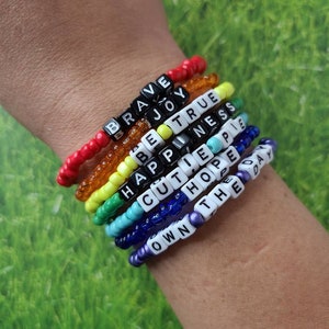 Pode incluir: Um conjunto de oito pulseiras de contas coloridas com palavras escritas em contas de letras brancas. As pulseiras são vermelhas, laranja, amarelas, verdes, azuis, roxas e brancas. As palavras nas pulseiras são "Brave", "Joy", "Be True", "Happiness", "Cutie Pie", "Hope", "Own The Day".
