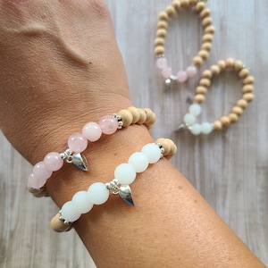 May include: Two bracelets with wooden beads and gemstone accents. One bracelet has pink gemstone beads and the other has white gemstone beads. Both bracelets have a silver charm in the shape of a leaf.