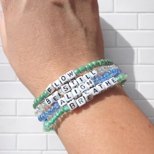 May include: Four beaded bracelets in shades of green and blue, with white letter beads spelling out the words "FLOW", "BE STILL", "ALIGN", and "BREATHE". The bracelets are stacked on a wrist.