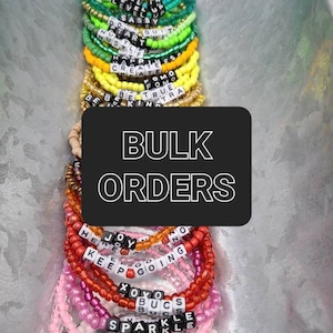May include: A close-up of a variety of colorful beaded bracelets with letter beads spelling out words and phrases like "Show Up", "Keep Going", "Sparkle", and "Angel". The bracelets are arranged in a pile on a silver surface. The text "BULK ORDERS" is displayed in a black box in the center of the image.