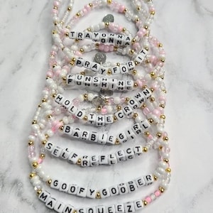 May include: A collection of beaded bracelets featuring white, pink, and gold-toned beads. Each bracelet includes white letter beads spelling phrases such as TRAYONNA, PRAY FOR IT, FUNSHINE, HOLD THE CROWN, BARBIE GIRL, SELF REFLECT, GOOFY GOOBER, and MAIN SQUEEZE.
