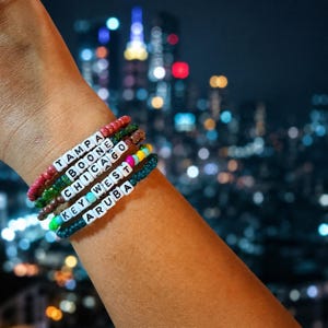 Puede incluir: Una pila de cinco pulseras de cuentas con cuentas cuadradas blancas con letras que deletrean TAMPA, BOONE, CHICAGO, KEY WEST y ARUBA. Las pulseras están hechas de pequeñas cuentas de vidrio de colores en tonos rojo, verde, marrón, amarillo y azul.