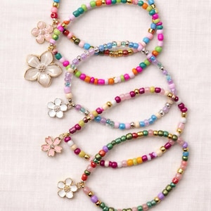 May include: Colorful beaded bracelets with flower charms. The bracelets feature a variety of small, round beads in shades of pink, green, blue, yellow, and gold. Each bracelet has a flower charm in white or pink.