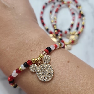 Puede incluir: Un brazalete de cuentas rojas, blancas y negras con un dije de Mickey Mouse dorado cubierto de diamantes de imitación transparentes.