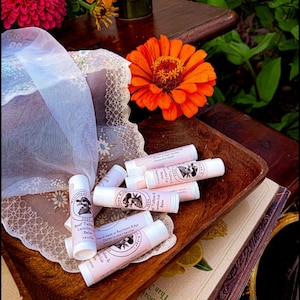 May include: A collection of cylindrical lip balm tubes with light pink labels featuring the text "Beef Tallow & Lanolin Lip Balm" and a circular logo with a bird illustration. The tubes are arranged in a wooden bowl with a white lace fabric.
