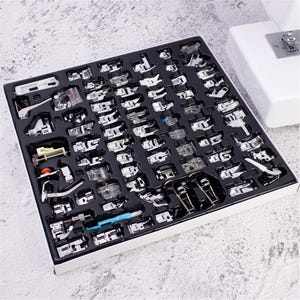 Puede incluir: Un conjunto completo de varios prensatelas para máquina de coser de metal y plástico, organizados en una bandeja moldeada negra. La colección incluye diversos accesorios para diferentes técnicas de costura, con componentes de metal plateado y una herramienta de plástico azul.