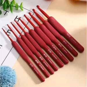 May include: A set of nine red crochet hooks with ergonomic handles, arranged diagonally. Each hook is clearly labeled with its size, ranging from 2/0 (2.0mm) to 10/0 (6.0mm), printed in white text on the handle of each individual tool.