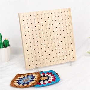 May include: A light-colored wooden pegboard with numerous evenly spaced holes. The pegboard is propped up by clear plastic stands. Two colorful, crocheted squares in shades of blue, brown, and red are in front of the board.
