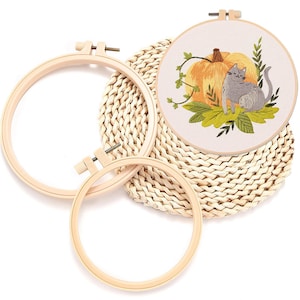Puede incluir: Un conjunto de bordado con aros de madera y una pieza terminada. El diseño bordado incluye un gato gris, una calabaza y hojas. Los aros son de madera clara y el fondo es una estera tejida.
