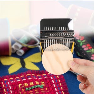 May include: A black and silver weaving tool with a wooden circular piece. The tool has multiple metal prongs and a yellow band. The background includes spools of thread and a colourful textile with a butterfly design.
