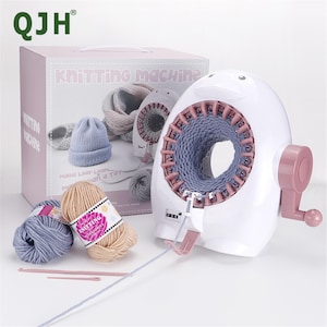 Könnte beinhalten: Eine weiße Strickmaschine mit rosa Griff und Akzenten, neben einer Box mit der Aufschrift "Strickmaschine". Die Maschine hat ein blaues Strickrohr im Inneren. Zwei Wollknäuel und Strickzubehör befinden sich vor der Maschine.