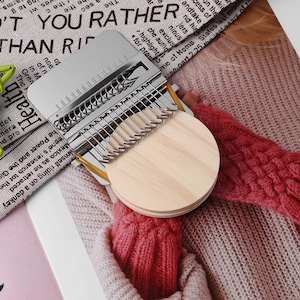 May include: A silver kalimba with a wooden soundboard and metal tines. The instrument features a circular wooden base. The kalimba is set against a backdrop of printed text and a pink knitted garment.