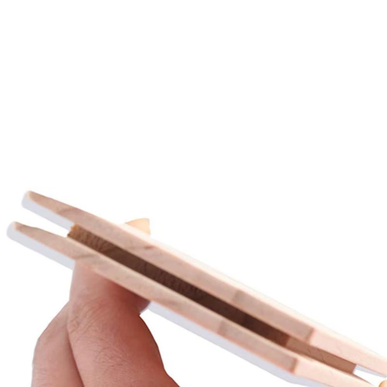 May include: A wooden clothes peg with a natural wood grain and white-painted edges. The peg is held in a hand, showcasing its design and construction. The image is set against a white background.