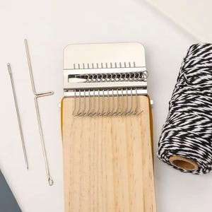 May include: A wooden weaving loom with a metal top and hooks, plus two metal needles and a spool of black and white yarn. The loom has a light wood finish, designed for crafting.