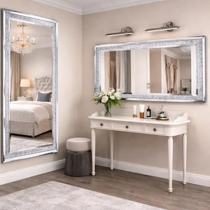 Può includere: Interno di una camera da letto con un grande specchio rettangolare con un bordo scintillante. Un tavolo da toeletta bianco con tre cassetti e uno sgabello rotondo sono di fronte a uno specchio rettangolare. Un lampadario si riflette nel grande specchio.