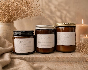 May include: Three glass jars of scented candles with black and gold lids, labeled "FRESH LINEN." The candles are on a stone surface, with a lit candle and dried flowers in the background. The jars have white labels with black text.