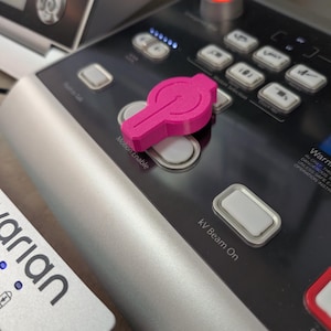 May include: A bright pink, 3D-printed button cover sits on a black control panel with silver trim. The panel has several buttons, including "Motion Enable" and "kV Beam On". A red light is illuminated above the button cover.