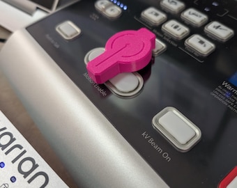 May include: A bright pink, 3D-printed button cover sits on a black control panel with silver trim. The panel has several buttons, including "Motion Enable" and "kV Beam On". A red light is illuminated above the button cover.