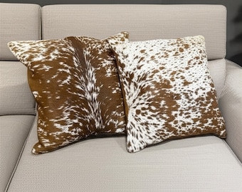 May include: Two decorative throw pillows with a cowhide pattern. The pillows feature a brown and white spotted design, with a natural texture. They are rectangular in shape and rest on a light-colored sofa.