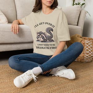 Puede incluir: Una camiseta beige de manga corta con el gráfico de un mapache gruñón tumbado. El texto sobre el mapache dice I'LL GET OVER IT y el texto debajo dice I JUST NEED TO BE DRAMATIC FIRST. La prenda se combina con unos pantalones vaqueros azules.