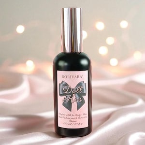 Peut inclure: Un flacon pulvérisateur en verre noir avec un bouchon argenté métallique et une étiquette rectangulaire rose clair. L'étiquette porte le texte SOLIVARA, Doll Kiss, Perfume Milk for Body & Hair, 100 ml / 3.4 fl oz et un motif de nœud noir.
