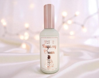 May include: A light green glass bottle with a rose gold cap and label. The label reads "SOLIVIA Raspberry Cream" with a dessert graphic. The background is a soft, blurred white with bokeh lights.